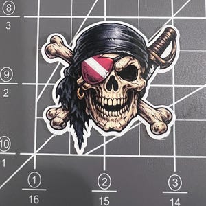 May include: A die-cut sticker featuring a detailed human skull wearing a black bandana and a red and white striped eye patch. Behind the skull are two crossed bones and a sword. The sticker is placed on a gray cutting mat with white grid lines.