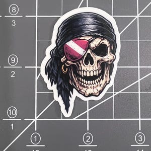 May include: A die-cut sticker featuring a detailed illustration of a human skull wearing a black bandana and a red and white striped eye patch. The skull has a gold hoop earring in one ear and a dark, textured appearance with visible teeth.