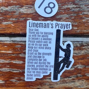 May include: A rectangular white sticker featuring the black text Lineman's Prayer followed by a prayer for safety and strength, accompanied by a black silhouette graphic of a worker climbing a utility pole, with a separate circular sticker numbered 18.