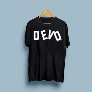 May include: Black t-shirt with the word "DEVO" in large, white, block letters. The shirt is hanging on a wooden hanger against a light blue background. The text is slightly angled.