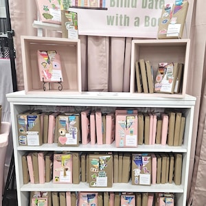 May include: A display of "Blind Date with a Book" products. Books are wrapped in brown paper and pink paper, decorated with stickers. The display includes a white bookshelf, pink boxes, and a banner that reads "Blind Date with a Book".