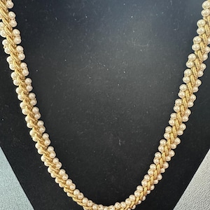 Puede incluir: Un collar dorado con un diseño trenzado. El collar presenta pequeñas cuentas blancas redondas intercaladas en la trenza dorada. El collar se muestra sobre un fondo negro.