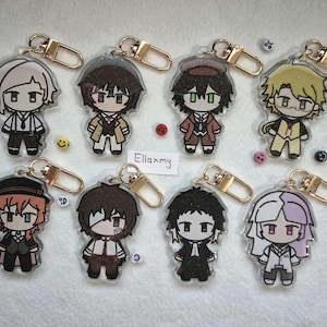 Peut inclure: Huit porte-clés en acrylique avec des personnages de style anime. Chaque porte-clés a un fermoir doré. Les personnages ont différentes couleurs de cheveux et tenues, avec des contours noirs et des accents colorés. De petites perles décoratives et un smiley sont éparpillés.