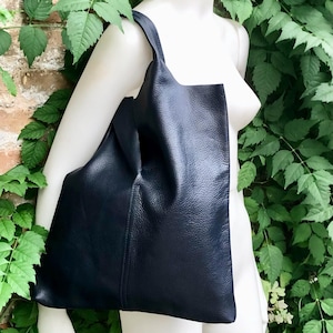 May include: A large, navy blue leather tote bag with a soft, textured appearance. The bag has a single shoulder strap and a spacious interior. The bag is displayed on a white mannequin, surrounded by green foliage.