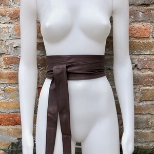 Op de afbeelding: Een bruine leren riem met een lange, vloeiende stropdas. De riem is om een wit mannequin gebonden.