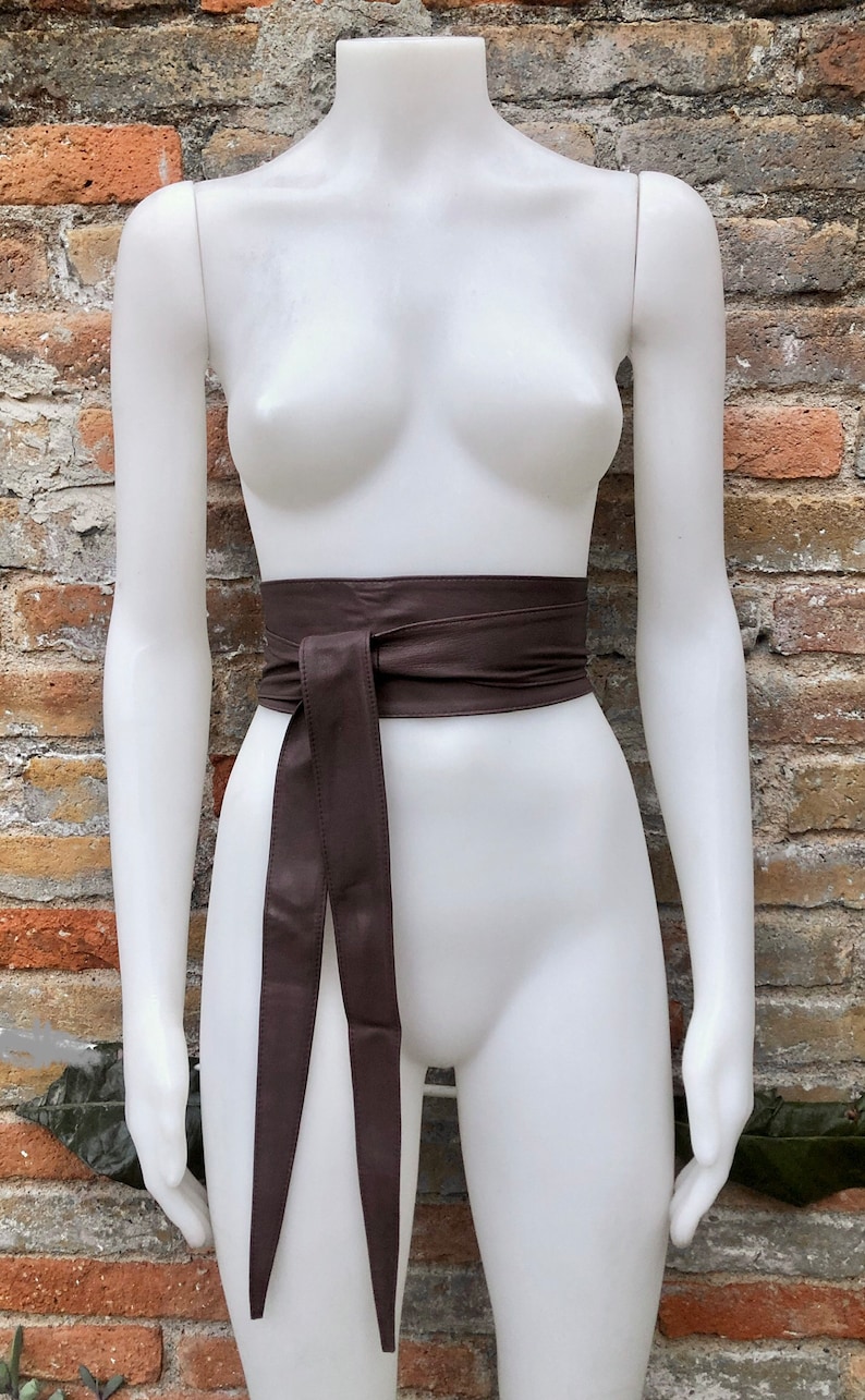 Op de afbeelding: Een bruine leren riem met een lange, vloeiende stropdas. De riem is om een wit mannequin gebonden.