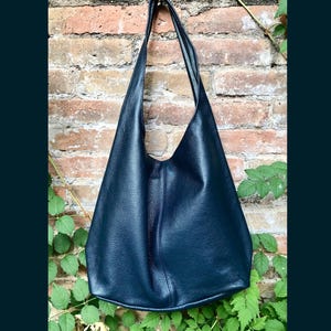 Op de afbeelding: Een donkerblauwe leren draagtas met een getextureerd oppervlak. De tas heeft een lange riem en hangt tegen een bakstenen muur met groen gebladerte.