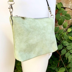Könnte beinhalten: Eine hellgrüne Wildleder-Umhängetasche mit silbernem Reißverschluss und Beschlägen. Die Tasche hat eine rechteckige Form und einen langen, verstellbaren Riemen. Die Tasche wird vor einem Hintergrund aus grünen Blättern und einer Backsteinmauer gezeigt.