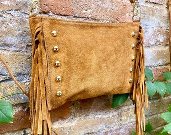 May include: A tan suede crossbody bag with fringe detailing and gold-colored stud accents. The bag has a long, adjustable strap and a rectangular shape. The fringe hangs from the sides and bottom of the bag, adding a bohemian flair.