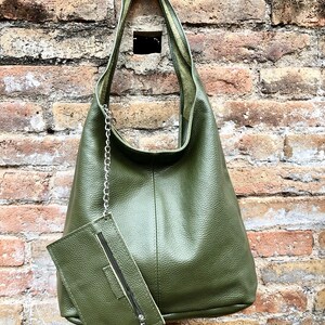 May include: Olive green leather shoulder bag with a matching zippered pouch attached by a silver chain. The bag has a textured surface and a simple, elegant design. The background is a weathered brick wall.