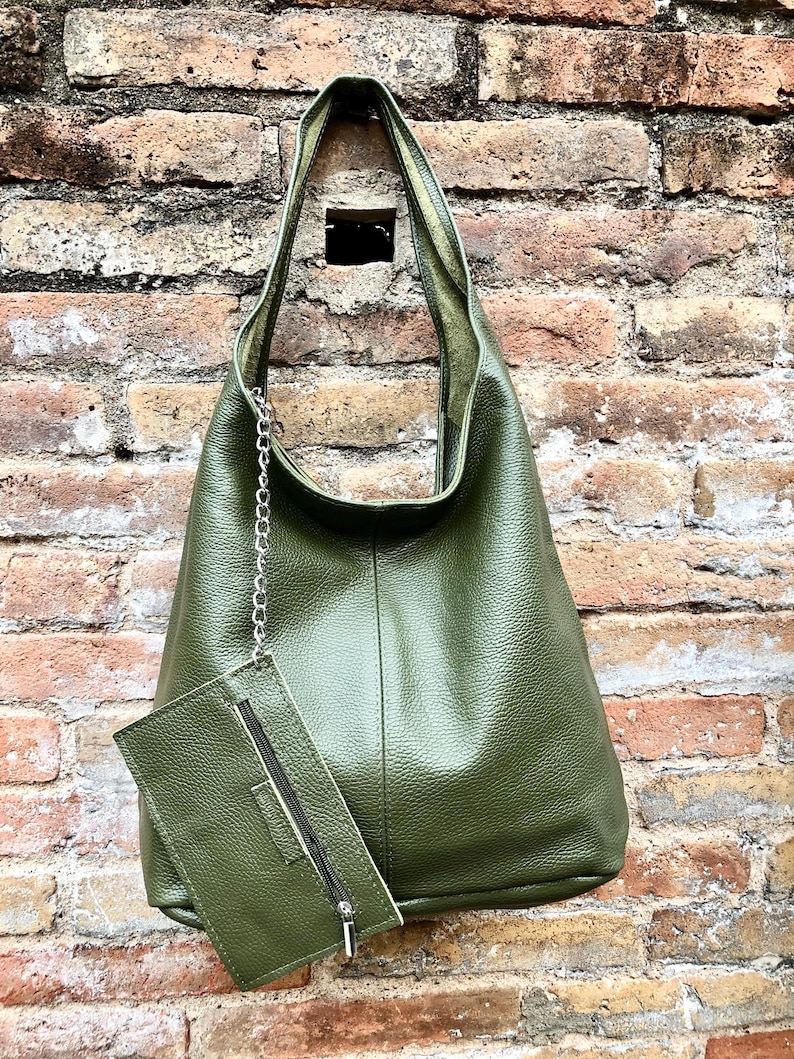 May include: Olive green leather shoulder bag with a matching zippered pouch attached by a silver chain. The bag has a textured surface and a simple, elegant design. The background is a weathered brick wall.