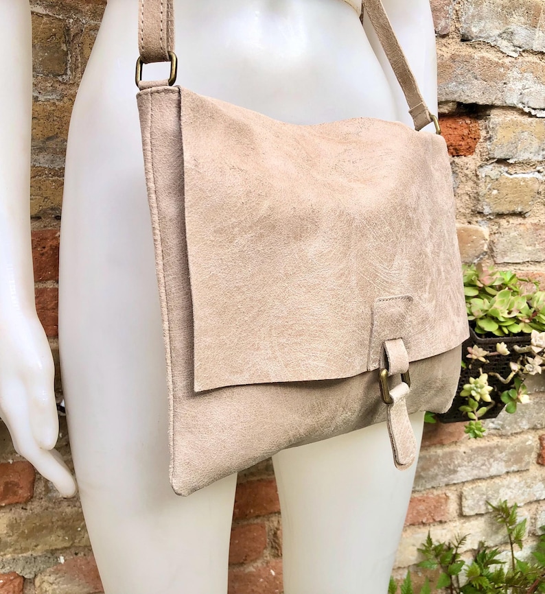 May include: A light beige leather shoulder bag with a flap closure and a buckle detail. The bag has a long strap and is made of a soft, textured leather.