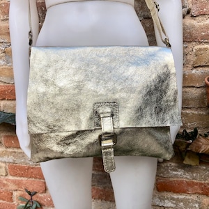 May include: A silver metallic leather crossbody bag with a buckle closure. The bag has a flap that folds over and is secured with the buckle.