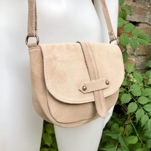 May include: A beige suede crossbody bag with a flap closure and a strap with gold-toned accents.