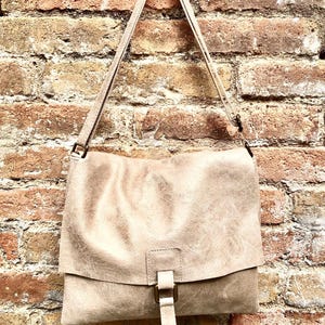 May include: A light brown leather shoulder bag with a flap closure and a buckle. The bag is hanging on a brick wall.