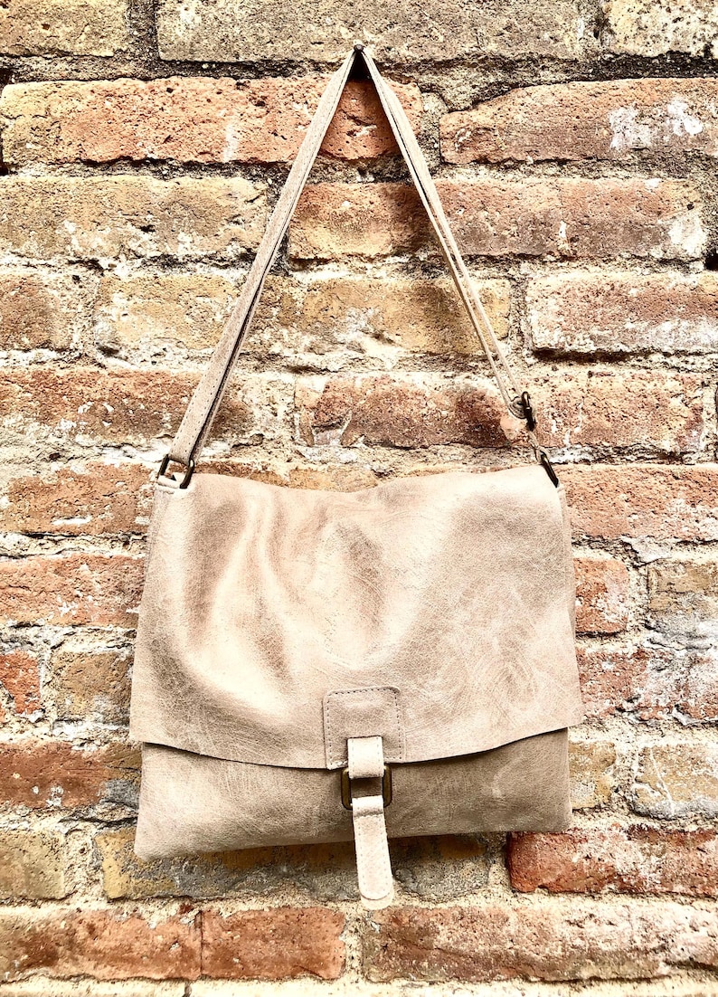 May include: A light brown leather shoulder bag with a flap closure and a buckle. The bag is hanging on a brick wall.