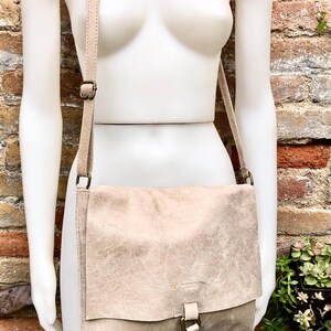May include: A light beige leather crossbody bag with a flap closure and a buckle. The bag has a long strap and is made of a textured leather.