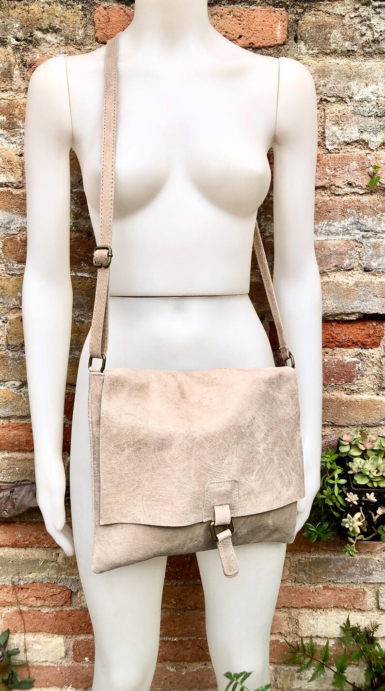 May include: A light beige leather crossbody bag with a flap closure and a buckle. The bag has a long strap and is made of a textured leather.