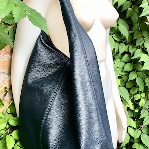 May include: A large, dark blue leather shoulder bag with a textured surface. The bag has a single strap and a spacious, open design. The bag is displayed against a background of green foliage and a brick wall.