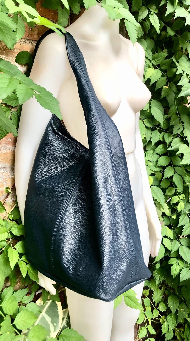 May include: A large, dark blue leather shoulder bag with a textured surface. The bag has a single strap and a spacious, open design. The bag is displayed against a background of green foliage and a brick wall.