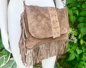 May include: A brown suede crossbody bag with a flap closure and fringe detailing. The bag features a strap and is adorned with small, gold-colored studs. The bag is displayed on a mannequin.