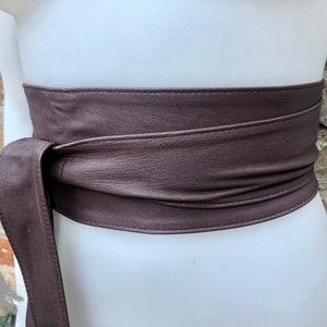 Op de afbeelding: Een bruine leren riem met een lange strik. De riem is om een wit mannequin torso gebonden.