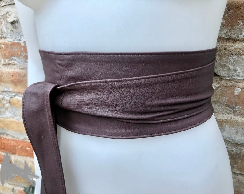 Op de afbeelding: Een bruine leren riem met een lange strik. De riem is om een wit mannequin torso gebonden.