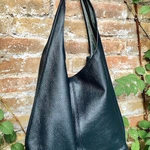 May include: A dark teal leather tote bag with a textured surface. The bag has a long strap and a large, open design. It hangs against a brick wall with green foliage.