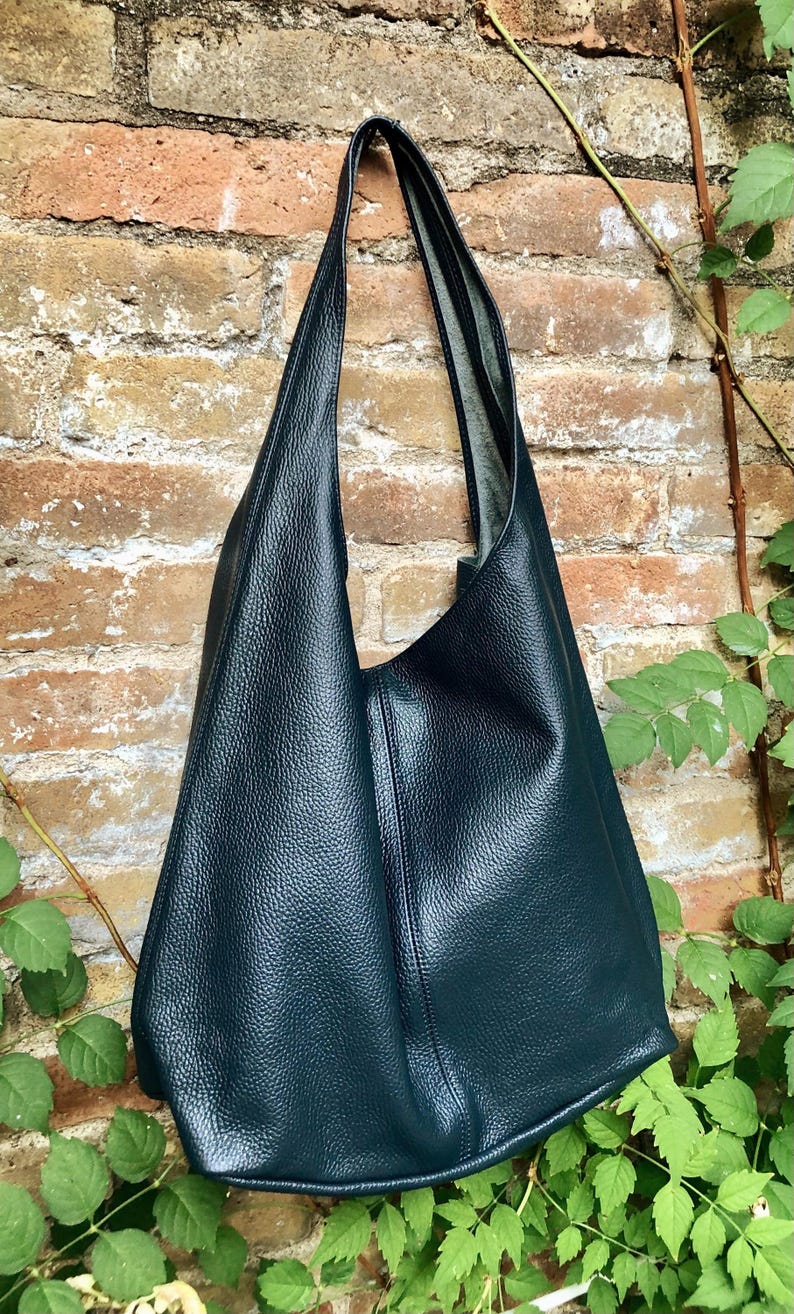 May include: A dark teal leather tote bag with a textured surface. The bag has a long strap and a large, open design. It hangs against a brick wall with green foliage.