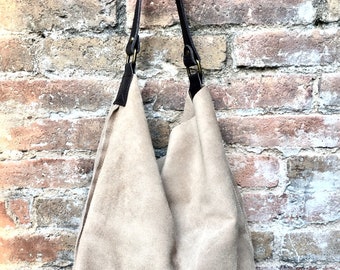 May include: A beige suede tote bag with brown leather straps and gold buckles. The bag is hanging on a brick wall.