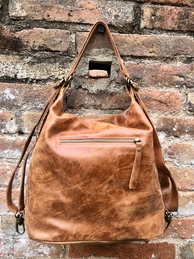 Op de afbeelding: Een bruine leren rugzak met een ritsvak en verstelbare schouderbanden. De tas heeft een verweerde, vintage look.