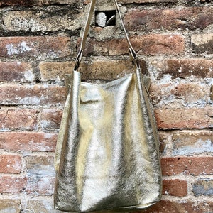 May include: A gold-coloured leather tote bag with a long strap hanging from a brick wall.