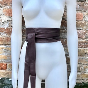 Op de afbeelding: Een bruine leren riem met een lange, dunne stropdas. De riem is om de taille van een wit mannequin gebonden.