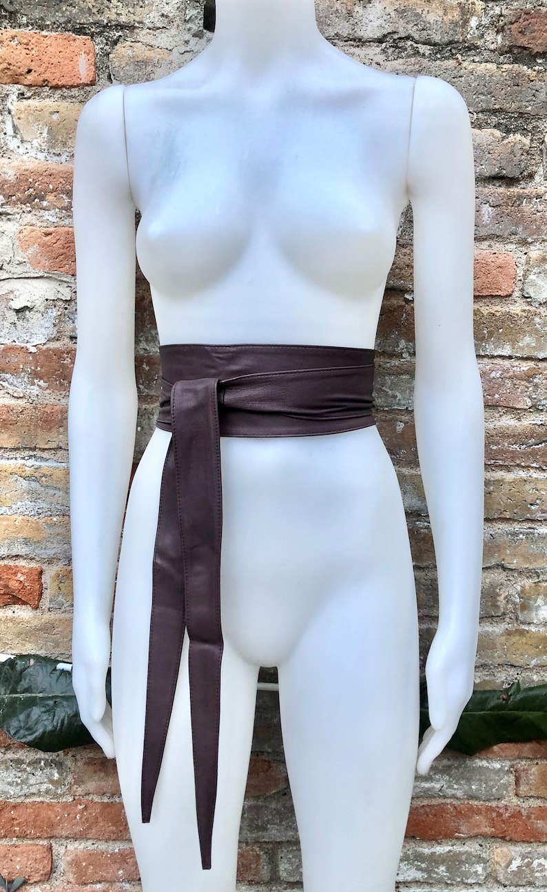 Op de afbeelding: Een bruine leren riem met een lange, dunne stropdas. De riem is om de taille van een wit mannequin gebonden.