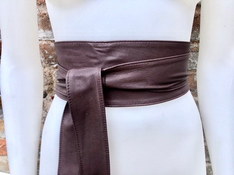 Op de afbeelding: Een bruine leren riem met een breed, plat ontwerp. De riem is aan de voorkant in een knoop geknoopt en heeft een gladde, gepolijste afwerking.