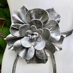 Puede incluir: Un cinturón de flores de cuero plateado. El cinturón presenta un diseño de flor grande y texturizado con múltiples pétalos. La correa del cinturón también es plateada y parece estar hecha del mismo material. El cinturón se exhibe en un maniquí blanco.