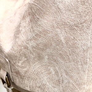 May include: Close-up of a light beige leather tote bag with a textured surface. The bag has a flap closure and a strap with a buckle.