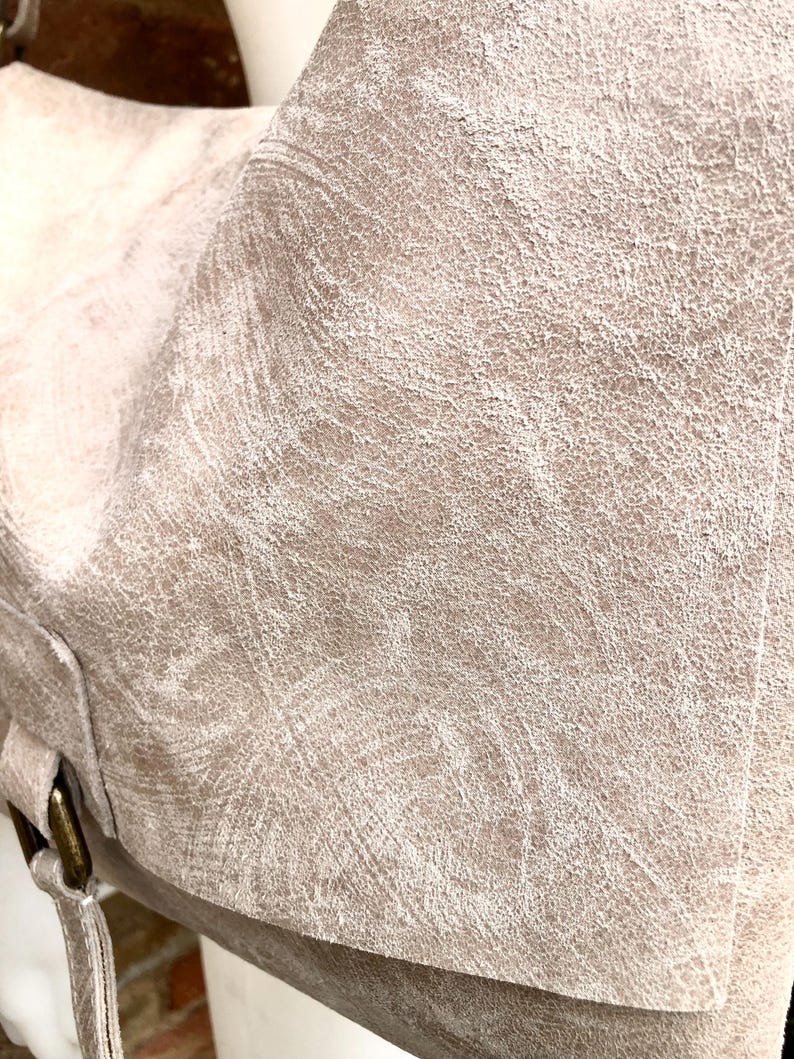 May include: Close-up of a light beige leather tote bag with a textured surface. The bag has a flap closure and a strap with a buckle.