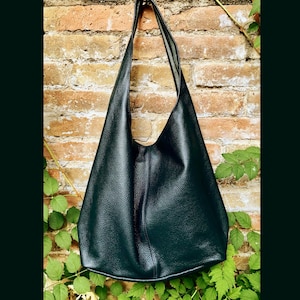 May include: A dark green leather shoulder bag with a long strap. The bag has a simple, minimalist design and is set against a brick wall with green foliage.