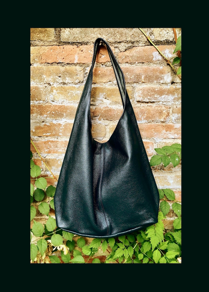 May include: A dark green leather shoulder bag with a long strap. The bag has a simple, minimalist design and is set against a brick wall with green foliage.
