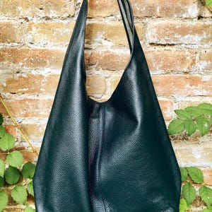 May include: A dark teal leather shoulder bag with a textured surface. The bag has a long strap and a large, open design. The bag is hanging against a brick wall with green foliage.