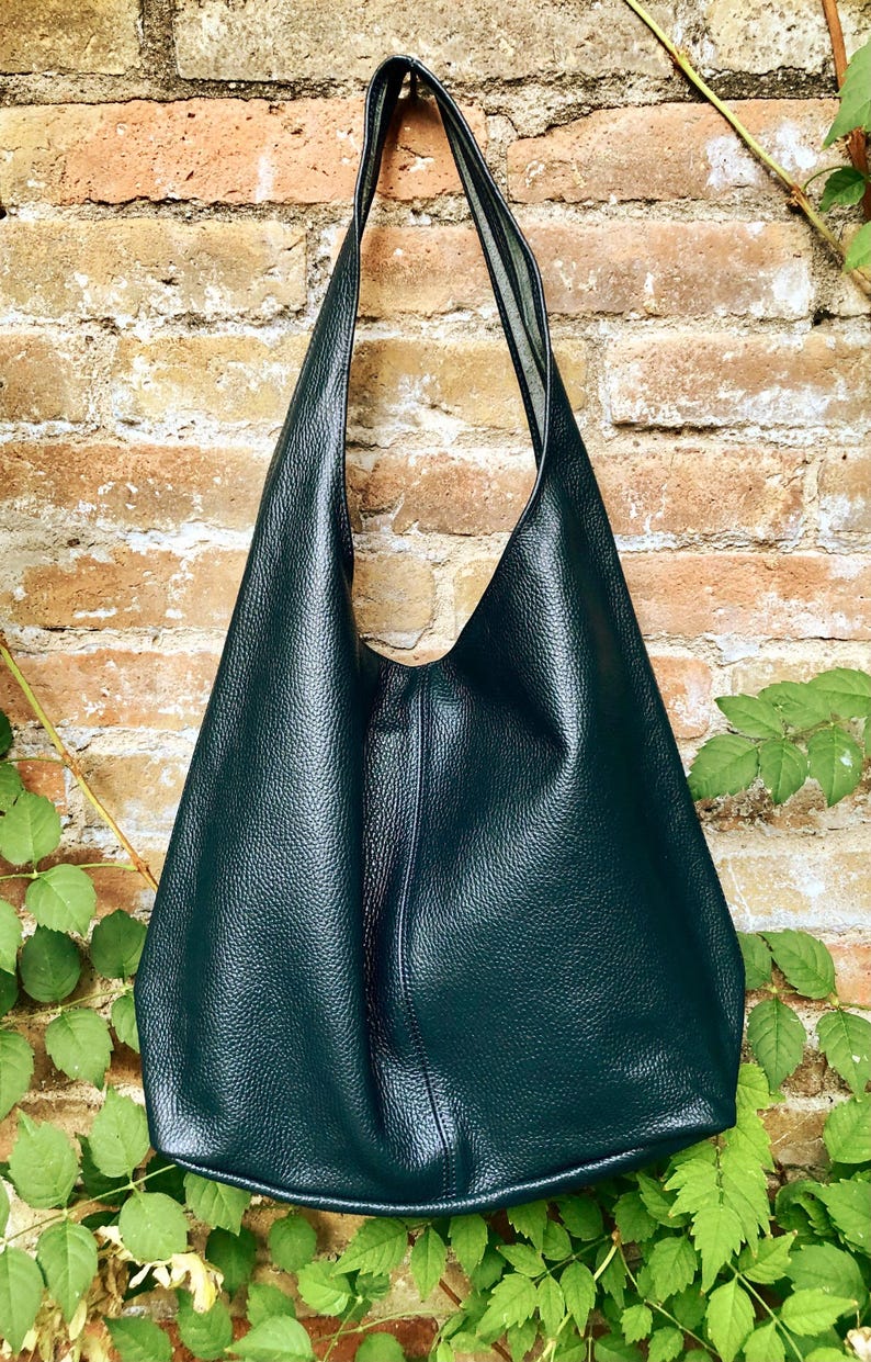 May include: A dark teal leather shoulder bag with a textured surface. The bag has a long strap and a large, open design. The bag is hanging against a brick wall with green foliage.