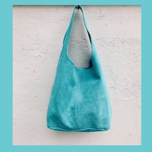 May include: A turquoise blue suede tote bag hanging on a white wall. The bag has a single strap and a slouchy, casual style.