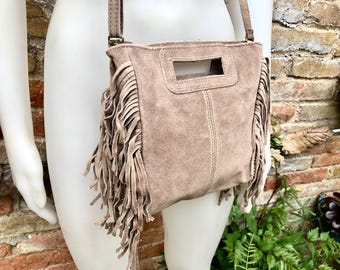 May include: A light brown suede crossbody bag with a fringe detail. The bag has a flap closure and a small rectangular cutout on the front.