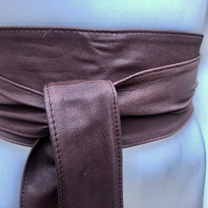 Op de afbeelding: Een bruine leren riem met een eenvoudig ontwerp. De riem is aan de voorkant in een knoop geknoopt.