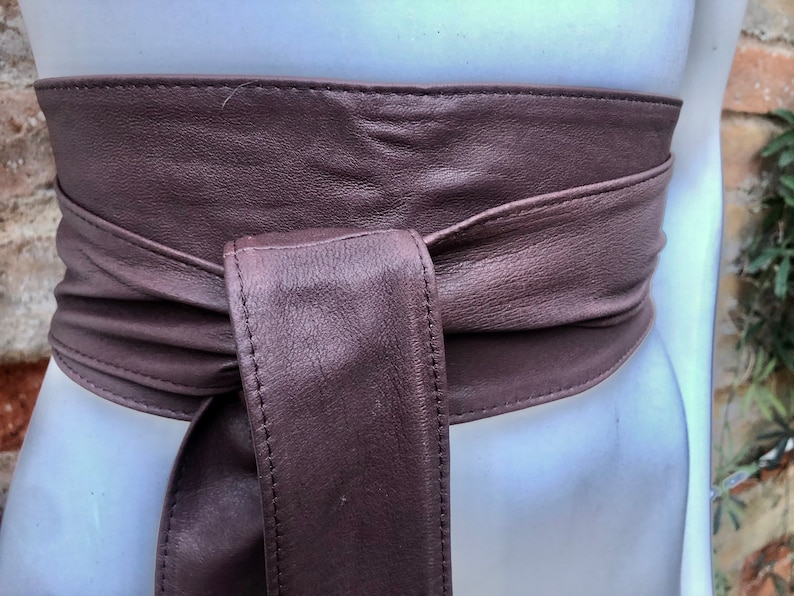 Op de afbeelding: Een bruine leren riem met een eenvoudig ontwerp. De riem is aan de voorkant in een knoop geknoopt.