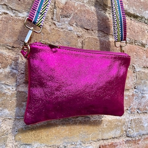 May include: A metallic fuchsia crossbody bag with a colourful striped strap. The bag has a rectangular shape with a gold zip and hardware. The strap features pink, green, yellow, and blue stripes.