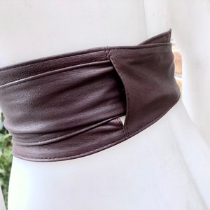 Op de afbeelding: Een bruine leren riem met een uniek ontwerp met een uitsparing in het midden. De riem is dubbelgevouwen en vastgemaakt met een gesp, wat een stijlvolle en moderne uitstraling cre&euml;ert.