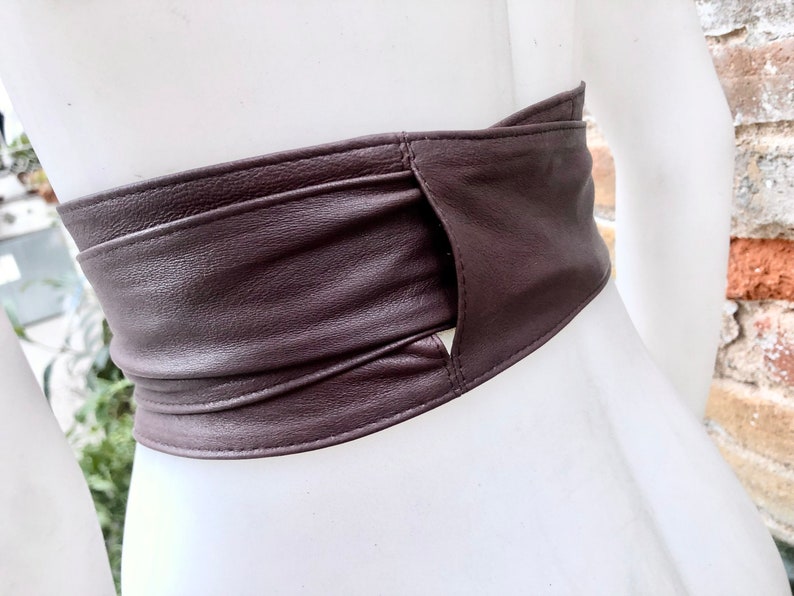Op de afbeelding: Een bruine leren riem met een uniek ontwerp met een uitsparing in het midden. De riem is dubbelgevouwen en vastgemaakt met een gesp, wat een stijlvolle en moderne uitstraling cre&euml;ert.