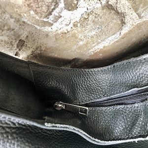 May include: Close-up of a dark gray leather bag with a textured surface. The bag has a silver zipper and a contrasting white lining. The image shows the inside of the bag, with a glimpse of the opening.